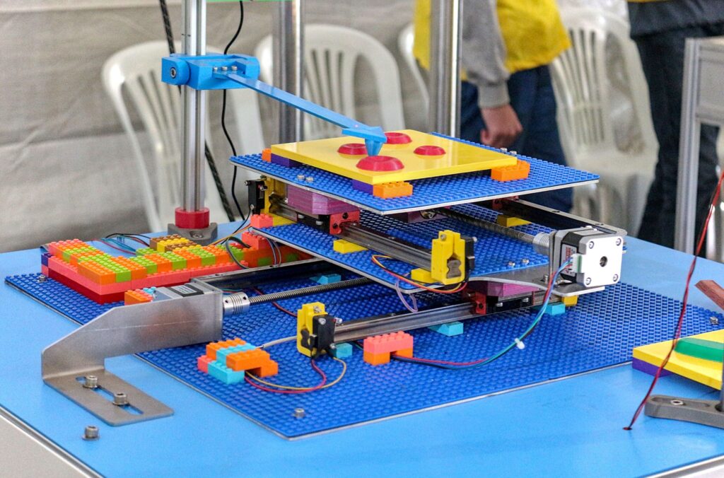 1. Como uma vitrola nos ensina sobre o mundo submicroscópico. Nesta fotografia pode-se identificar um aparato feito com peças de lego e alguns componentes metálicos e eletrônicos. Ela têm uma estrutura com várias camadas de placas, presas com parafusos e espaçadas entre si, formando o sistema que pode se mover tanto na vertical quanto na horizontal. Na parte de cima há um braço mecânico com uma ponta que tateia a superfície de uma amostra. Isso representa o funcionamento de um microscópio de força atômica. Tal equipamento tateia a superfície de uma amostra, assim se tornando capaz de "enxergar" as nuances de tal superfície, descobrindo detalhes muito além do que o olho humano consegue enxergar.